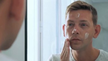 Kafkasyalı yakışıklı genç adamın banyoda durup aynaya baktıktan sonra yüzüne nemlendirici krem sürüşünün yansıması. Günlük rutin erkek hijyenik güzellik prosedürü.
