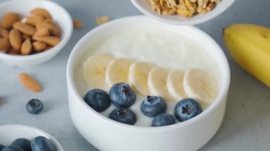 Kadın eli, Yunan yoğurdunun içine yulaf ezmesi, muz ve sağlıklı kahvaltı hazırlamak için gri arka planda yaban mersini ekliyor. Diyet yemek.