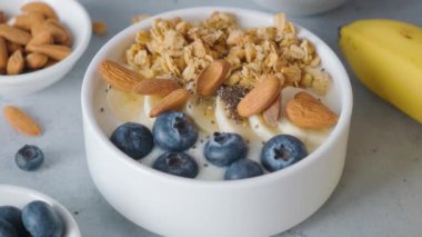 Kadın eli, sağlıklı kahvaltı hazırlamak için arka planda yulaf, muz, yabanmersini ve badem içeren Yunan yoğurdu kasesine chia tohumu ekliyor. Diyet yemek.