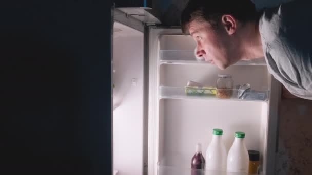 夜のオープン冷蔵庫の検索食品で暗いキッチンの自宅で白人の空腹の男の若い男性 冷蔵庫からサラダレタスを取って夜の飢餓と食事に不満を持つ男 不眠症の概念 — ストック動画