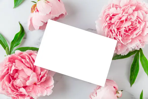 Blank greeting card in frame made of pink peony flowers on white marble background. Wedding invitation. Mock up. Flat lay. Womens day, Valentines day, Mothers day layout.