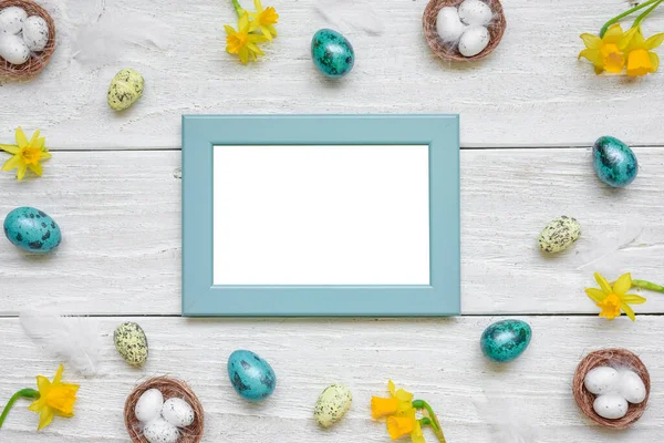 Easter picture photo frame with spring yellow Narcissus flowers and easter colorful eggs on white wooden background. Mock up. Flat lay. Top view with copy space.