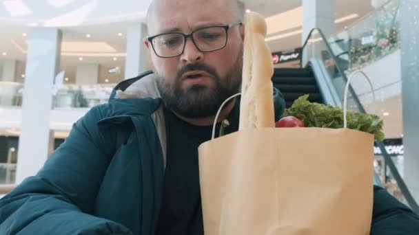 食べ物がいっぱいのショッピングペーパーバッグが付いている失望したプラスサイズの男性は スーパーマーケットのエスカレーターの販売紙レシートを見ます 食糧インフレとショッピングモールの価格の上昇にショックを受けた男性 — ストック動画