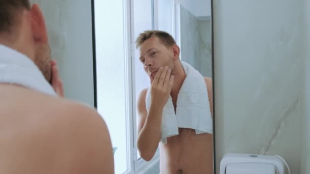 若いハンサムな若い男はタオルでトイレに立って鏡を見ている顔に彼のブリストルを浮かべます 毎日の朝のルーチン男性衛生美容手順のための準備 — ストック動画
