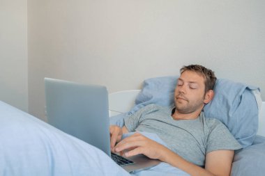 Genç adam sabahları bir otel odasında yatarken dizüstü bilgisayarla çalışıyor. Bilgisayarda daktilo eden serbest çalışan bir erkek. Yatak odasında iş, sosyal ağlar, sörf, internette gezinme, uzaktan çalışma.