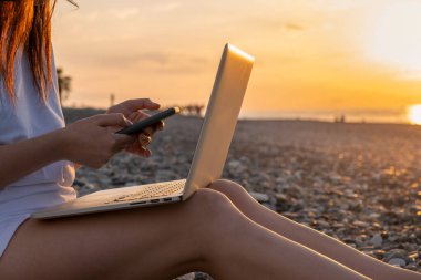 Gün batımında sahilde dizüstü bilgisayar ve cep telefonuyla çalışan bir kadına yakın çekim. Serbest çalışan bir kadın uzaktan akıllı telefon ve bilgisayar kullanarak çevrimiçi çalışıyor. Tatilde uzaktan çalışma
