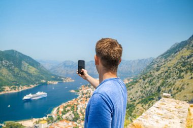Akıllı telefondan fotoğraf çeken turist Kotor Körfezi 'nin yukarısındaki Lovcen Dağı ve Karadağ' ın eski kenti üzerinde duruyor. Erkek gezgin, yazın Adriyatik fiyortunun güzel manzarasını cep telefonuyla çekiyor.