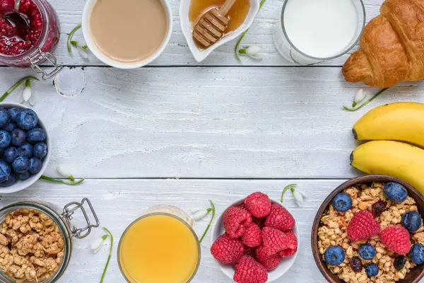 Healthy breakfast buffet with various morning food and spring flowers. Frame made of granola, coffee, fruit, berries, milk, orange juice and honey on white wooden table. Top view with copy space.