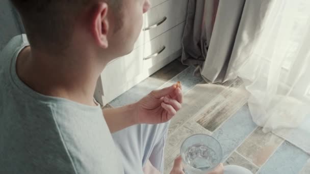 オメガ3のカプセルを飲む男 自宅のベッドに座っている水のガラスとビタミンD 魚油と栄養補助食品 健康支援と治療のコンセプト バックビュー — ストック動画