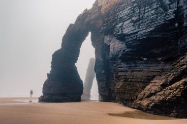 Tourist walking along natural arch on Cathedrals beach in Galicia, Spainn. Man silhouette in foggy landscape with Playa de Las Catedrales Catedrais beach in Ribadeo, Lugo on Cantabrian coast. Moody clipart