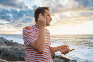 Deniz kıyısında kulaklık takan ve müzik dinleyen akıllı telefonlu neşeli beyaz bir genç. Yakışıklı erkek gün batımında plajda kulaklıklı cep telefonu kullanıyor..