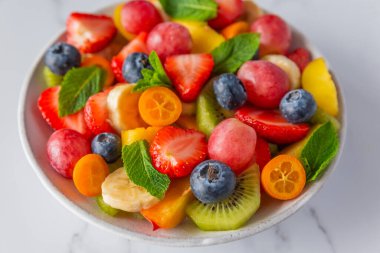 Beyaz mermer arka planda bir kasede yaz meyve salatası. Kahvaltı için sağlıklı diyet yemeği. Diyet öğle yemeği için meyve, böğürtlen ve nane.