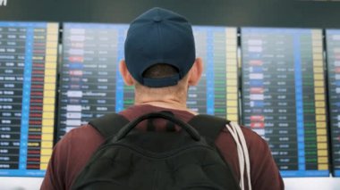 Sırt çantalı bir adam havaalanındaki uçuş bilgilerini kontrol ediyor, yolculuğuna hazırlanıyor. Havaalanı terminalindeki erkek turistin arka görüntüsü uçak çizelgesine bakıyor. Seyahat ve