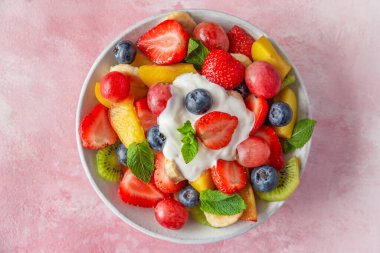 Yoğurtla kaplanmış taze meyve salatası ve nane yapraklarıyla süslenmiş çilek, yabanmersini, kivi, üzüm, muz ve pembe arka planda şeftali. Üst Manzara. Sağlıklı yaz yemekleri.. 
