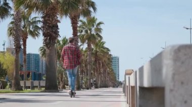 Arkadaki görüntüde, erkek turist, deniz kenarında palmiye ağaçları ve yeşilliklerle elektrikli scooter kullanıyor. Erkek, açık havada e-scooter kullanıyor. Yeşil enerji ve sürdürülebilir yaşam tarzı kavramı.