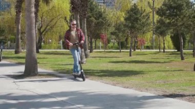 Güneş gözlüklü bir adam kaldırımlı bir yolda elektrikli scooterla palmiye ağaçları ve yeşillikler arasındaki manzaralı bir parkın içinden geçiyor. Erkek, açık havada e-scooter kullanıyor. Yeşil enerji ve sürdürülebilir yaşam tarzı kavramı.