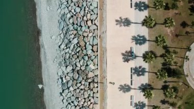 Güneşli bir günde, kıyı şeridi manzarası, palmiye ağaçları ve turkuaz sular boyunca kayalık bir kıyı şeridi. Deniz kıyısındaki gezinti güvertesi boyunca gölgeli tropik yaz palmiyeleri. Üst Manzara. Soyut