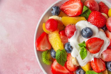 Sağlıklı taze meyve salatasını pembe arka planda yoğurt kasesiyle kapatın. Kopyalama alanı olan üst görünüm. Kahvaltı için sağlıklı yaz yemekleri. Vejetaryen diyeti öğle yemeği için karışık tropikal meyve..