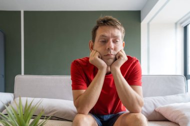 Kırmızı tişörtlü sıkkın adam, çenesi ellerinin üzerinde kanepede oturuyor. Modern oturma odasındaki kanepede oturan yorgun erkek. Stres, depresyon melankoli ve akıl sağlığı konsepti