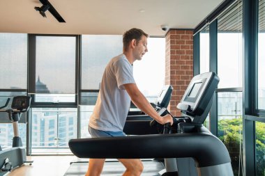 Spor antrenmanı için ısınan erkek sporcu ve şehir manzaralı modern spor salonunda koşu bandında yürüyor. Adam rezidans binasındaki fitness stüdyosunda kardiyo eğitimi alıyor. Sağlık, sağlık, sağlık.
