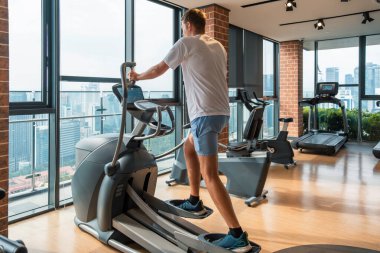 Modern spor salonunda şehir manzaralı eliptik bir antrenörde çalışan bir adam. Erkek, apartmandaki fitness stüdyosunda eliptik makinede kardiyo eğitimi alıyor. Fitness, sağlık, sağlıklı yaşam tarzı