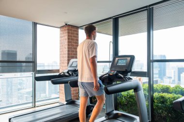 Spor antrenmanı için ısınan erkek sporcu ve şehir manzaralı modern spor salonunda koşu bandında yürüyor. Adam rezidans binasındaki fitness stüdyosunda kardiyo eğitimi alıyor. Sağlık, sağlık, sağlık.