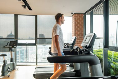 Spor antrenmanı için ısınan erkek sporcu ve şehir manzaralı modern spor salonunda koşu bandında yürüyor. Adam rezidans binasındaki fitness stüdyosunda kardiyo eğitimi alıyor. Sağlık, sağlık, sağlık.