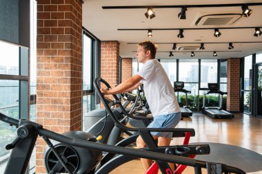 Modern spor salonunda şehir manzaralı eliptik bir antrenörde çalışan bir adam. Erkek, apartmandaki fitness stüdyosunda eliptik makinede kardiyo eğitimi alıyor. Fitness, sağlık, sağlıklı yaşam tarzı