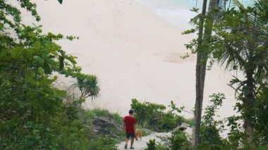Köpekli bir turist, turkuaz su okyanusuyla tropikal kumlu sahile giden taş merdivenlerden aşağı doğru yürür. Tayland 'ın Phuket adasındaki Özgürlük plajına yürüyen erkek turist..