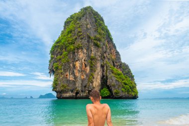 Tropikal kumsalda dinlenen turist Phra Nang Ko Rang Nok kayalık adasına bakıyor, Railay Bay, Krabi, Tayland. Erkek gezginin arkası turkuaz suyun kumlu sahilinde Andaman Denizi 'nde oturur.