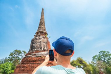 Tayland 'daki Ayutthaya tarihi parkında akıllı telefonuyla antik pagoda' nın fotoğrafını çeken bir turist. Erkek gezgin, arkeolojik sitede cep telefonuyla istorik Budist stupasını fotoğraflıyor.