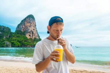 Turist, Tayland 'ın Krabi ilindeki kayalıklı tropikal kumlu Railay plajında plastik bardakta sarı mango smoothie içiyor. Erkek içecekleri yaz içecekleri Deniz kıyısında