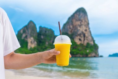 Adam, Tayland 'ın Krabi ilinde arka planda kayalıklar bulunan tropikal kumlu Railay plajında plastik bardakta sarı mango smoothie tutuyor. Deniz kıyısında yaz içkisi.