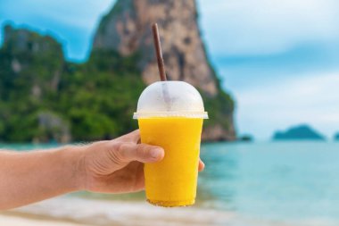 Tayland 'ın Krabi ilinde arka planda kayalıkları olan tropikal kumlu Railay plajında sarı mangolu smoothie tutan bir erkek. Erkekler ferahlatıcı yaz içkilerini plastik bardakta tutarlar.