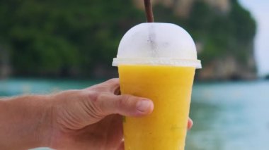 Tayland 'ın Krabi ilinde arka planda kayalıkları olan tropikal kumlu Railay plajında sarı mangolu smoothie tutan bir erkek. Erkekler ferahlatıcı yaz içkilerini plastik bardakta tutarlar.