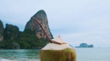 Tayland 'ın Krabi ilindeki kayalıklı tropikal kumlu Railay plajında taze hindistan cevizi içeceği tutan erkek. Deniz kıyısında bir pipetle ferahlatıcı bir yaz içeceği içen adam. Kapat..