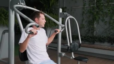 Modern balkonda göğüs pres makinesi üzerinde egzersiz yapan adam yeşil alana sahip. Erkek, rezistans binasının terasında spor aletleri eğitimi alıyor. Sağlık, sağlık, sağlık.