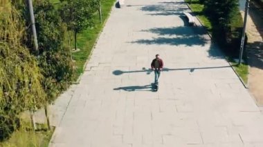 Güneşli bir yaz gününde, şehir parkında, köprüde elektrikli scooter süren bir adam. İHA 'dan yüksek açılı görüntü. Erkek, açık havada e-scooter kullanıyor. Çevre dostu nakliye ve seyahat. Sürdürülebilir yaşam tarzı