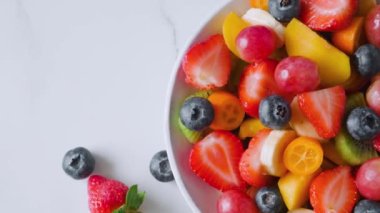 Beyaz mermer arka planda bir kasede taze meyve salatası. Üst Manzara. Kahvaltı için sağlıklı yemek. Karışık çilek, üzüm, muz, kivi, yaban mersini, şeftali, narenciye diyet öğle yemeği.