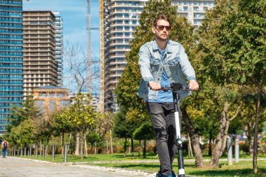 Güneşli yaz gününde şehir parkında e-scooter kullanan güneş gözlüklü bir adam. Erkek, elektrikli scooter kullanıyor ve açık havada eğleniyor. Çevre dostu şehir taşımacılığı ve seyahat. Sürdürülebilir yaşam tarzı
