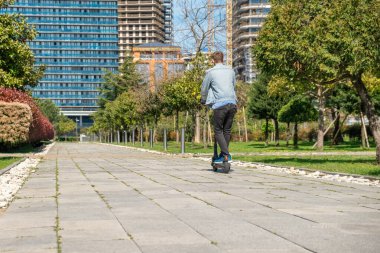 Güneşli yaz gününde, şehir parkında elektrikli scooter süren bir adamın arka görüntüsü. Erkek, e-scooter kullanıyor ve açık havada eğleniyor. Çevre dostu şehir taşımacılığı ve seyahat. Sürdürülebilir yaşam tarzı