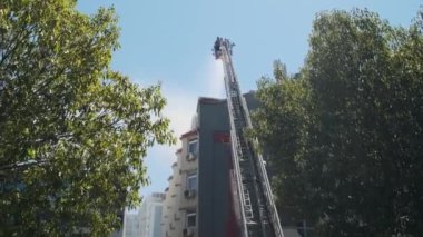 Yüksek platformdaki itfaiyecilerin arka görüntüsü yanan evi söndürmek için su hortumuyla donatılmış geniş merdiven kullanarak binanın çatısına duman püskürtüyor. İtfaiyeci yangını söndürüyor.