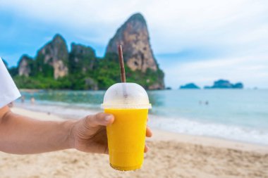 Tayland 'ın Krabi ilinde arka planda kayalıkları olan tropikal kumlu Railay plajında sarı mango smoothie tutan adam. Erkek el ferahlatıcı yaz içkisini plastik bardakta, kamışla birlikte tutar.