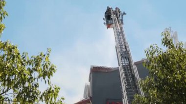 Yüksek platformdaki itfaiyecilerin arka görüntüsü yanan evi söndürmek için su hortumuyla donatılmış geniş merdiven kullanarak binanın çatısına duman püskürtüyor. İtfaiyeci yangını söndürüyor.