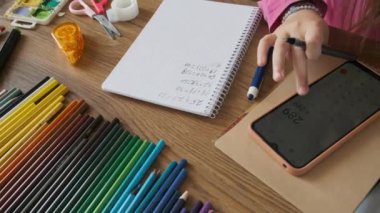 Matematik testi yapan, akıllı telefon kullanarak hesap yapan ve kağıt not defterine ahşap masadaki kağıtlarla yazan bir çocuk. Okul ödevi olarak kız el yazısı alıştırmaları. Okula dönüyorum, öğreniyorum.