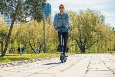 Güneşli yaz gününde şehir parkında e-scooter kullanan güneş gözlüklü bir adam. Erkek, elektrikli scooter kullanıyor ve açık havada eğleniyor. Çevre dostu şehir taşımacılığı ve seyahat. Sürdürülebilir yaşam tarzı