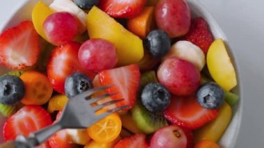 Beyaz mermer arka planda çatalı olan bir kasede taze meyve salatası yemek. Üst Manzara. Kahvaltı için sağlıklı yemek. Karışık çilek, üzüm, muz, kivi, yaban mersini, şeftali, diyet için narenciye.