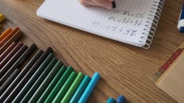 Çocuk, ahşap masadaki kırtasiyeyle defterde matematik alıştırmaları yapıyor. Okul ödevi olarak kız el yazısı matematik denklemleri. Okula dönüş, öğrenme, eğitim kavramı.