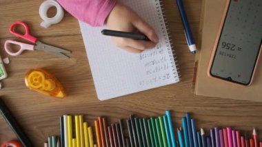 Çocuk, ahşap masadaki kırtasiyeyle denklemi çözmek için akıllı telefon hesap makinesi kullanarak defterde matematik testi yapıyor. Okul ödevi olarak kız el yazısı alıştırmaları. Üst Manzara. Öğrenme