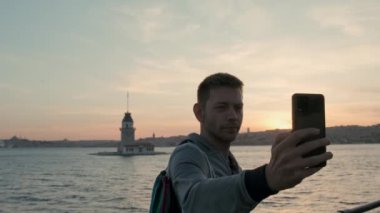 Erkek gezgin, cep telefonundaki kamerayla İstanbul 'un İstanbul Boğazı' ndaki Bakire Kulesi 'nde gün batımında selfie çekiyor. Kafkasyalı turist akıllı telefondan fotoğraf çekiyor. Yaz tatili